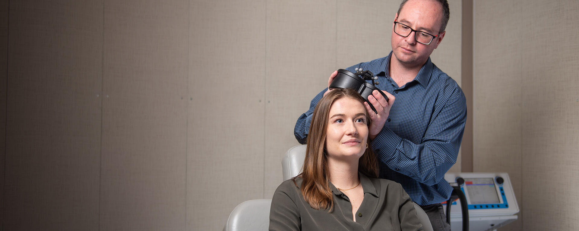 Dr. Alex McGirr demonstrates transcranial magnetic stimulation (TMS) on his graduate student, Jaeden Cole. McGirr is holding a small black device up against Jaeden's head.