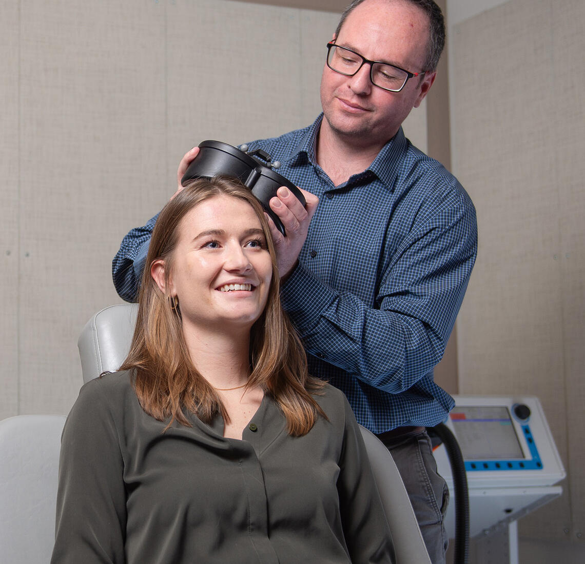 Dr. Alex McGirr demonstrates transcranial magnetic stimulation (TMS) on his graduate student, Jaeden Cole. McGirr is holding a small black device up against Jaeden's head.